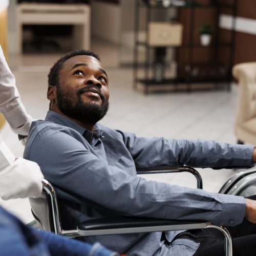 Happy young African American man using wheelchair during travel, receiving help and assistance in wheelchair-friendly and accessible hotel. People with mobility impairment and traveling